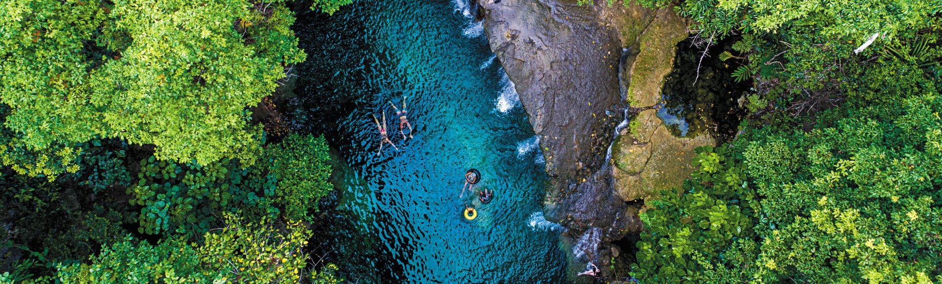 Aerial view of a vibrant turquoise natural pool surrounded by lush green foliage, with several people swimming and floating on tubes.