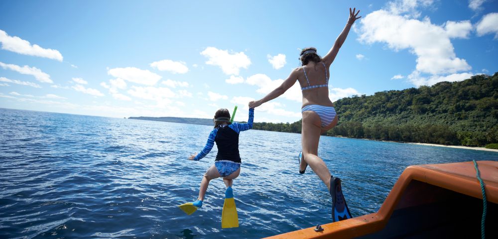 Two individuals, one wearing a blue rash guard and snorkeling gear, and another in a striped bikini, hold hands as they jump off the side of a boat into the clear blue ocean towards a tropical island shore.