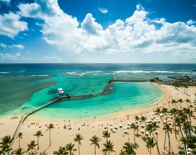Aerial view of a crowded tropical beach with turquoise water, palm trees, a pier with a boat, and a marina in the background under a partly cloudy blue sky.