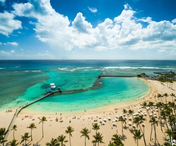 Aerial view of a crowded tropical beach with turquoise water, palm trees, a pier with a boat, and a marina in the background under a partly cloudy blue sky.