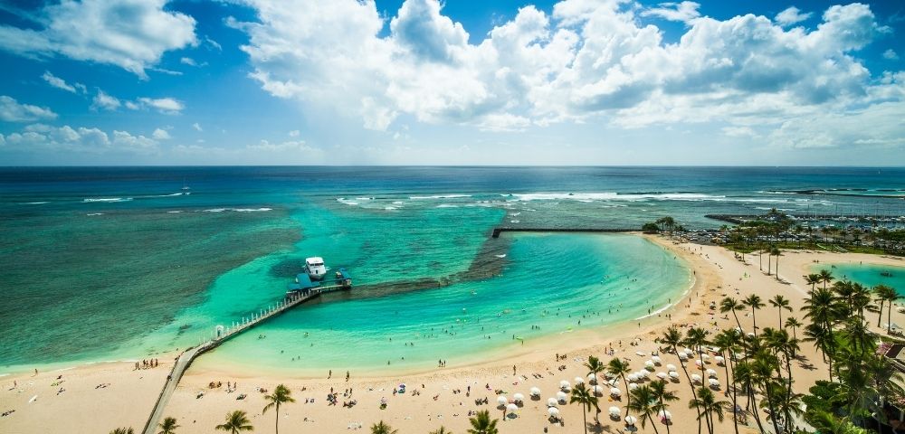 Aerial view of a crowded tropical beach with turquoise water, palm trees, a pier with a boat, and a marina in the background under a partly cloudy blue sky.