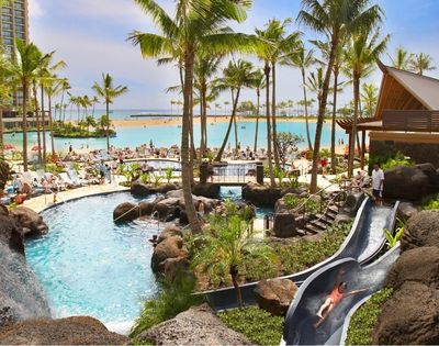 Resort with a large turquoise lagoon, swimming pools, a water slide, waterfalls, lush palm trees, and lounge chairs on a sandy beach.