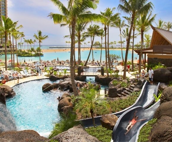 Resort with a large turquoise lagoon, swimming pools, a water slide, waterfalls, lush palm trees, and lounge chairs on a sandy beach.