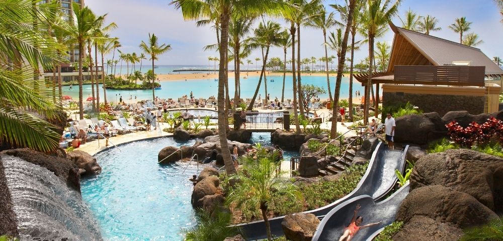 Resort with a large turquoise lagoon, swimming pools, a water slide, waterfalls, lush palm trees, and lounge chairs on a sandy beach.