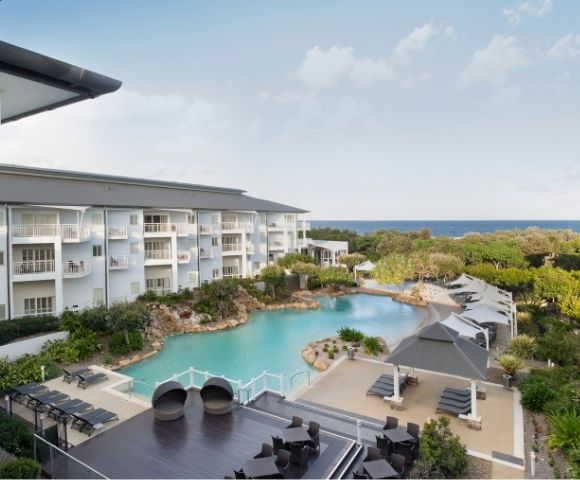 Oceanfront resort with a large pool, surrounded by white buildings, lounge chairs, umbrellas, and lush green landscaping under a partly cloudy sky.