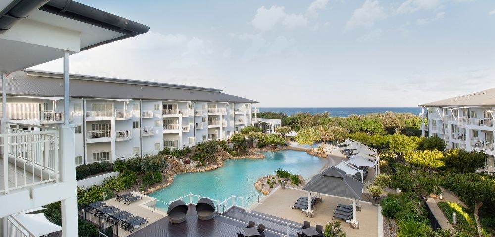 Oceanfront resort with a large pool, surrounded by white buildings, lounge chairs, umbrellas, and lush green landscaping under a partly cloudy sky.