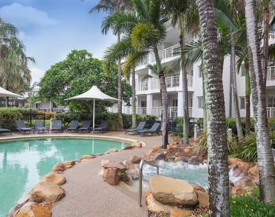 A tranquil swimming pool surrounded by palm trees and lounge chairs, with shaded cabanas nearby. The pool area is adjacent to a modern three-story building with balconies, set in a landscaped environment.