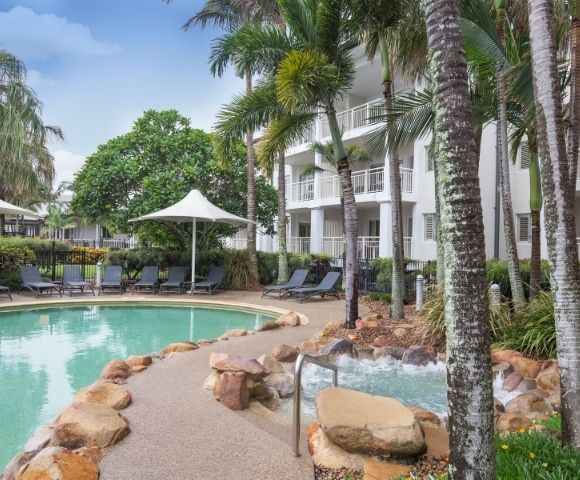A tranquil swimming pool surrounded by palm trees and lounge chairs, with shaded cabanas nearby. The pool area is adjacent to a modern three-story building with balconies, set in a landscaped environment.
