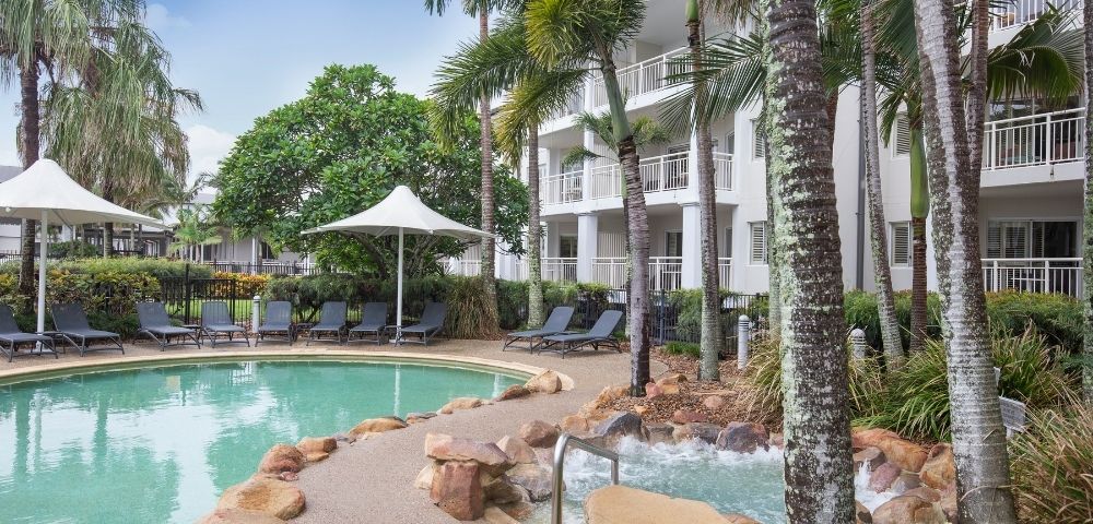 A tranquil swimming pool surrounded by palm trees and lounge chairs, with shaded cabanas nearby. The pool area is adjacent to a modern three-story building with balconies, set in a landscaped environment.