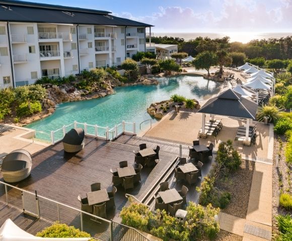 Aerial view of a beachfront hotel resort with a large lagoon-style swimming pool, surrounding lounge chairs, palm trees, and nearby buildings under clear daylight