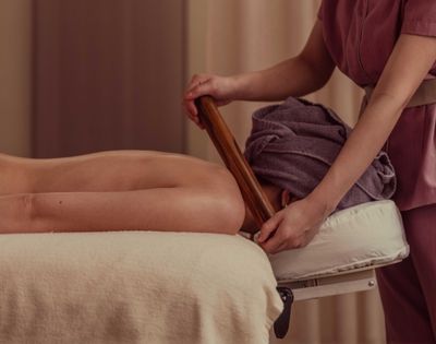 Person lying face down receiving a wooden stick massage from a therapist wearing a mauve uniform.