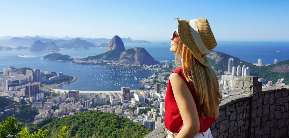 A woman in a hat and red top enjoys a scenic view over Rio de Janeiro, featuring Sugarloaf Mountain, blue sky, and calm sea, conveying serenity and wonder.