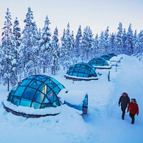 Snowy landscape featuring glass igloos nestled among frosty trees under a soft pink sky. Two people walk along a snow-covered path, conveying tranquility.