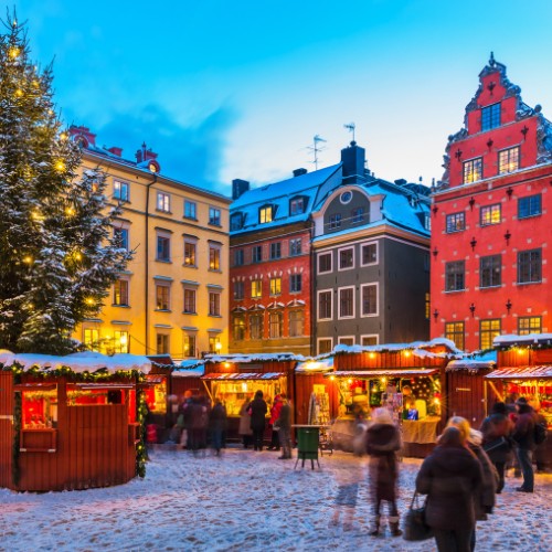 Snow-dusted Christmas market at twilight in a European town square. Festive stalls glow warmly under colorful historic buildings and a lit tree.