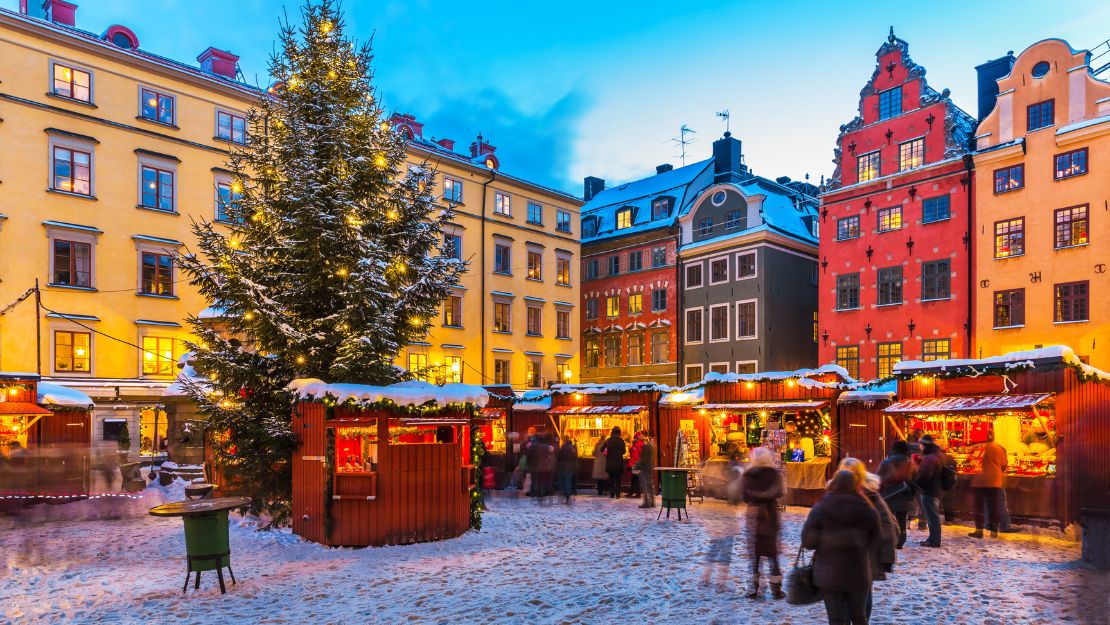Snow-dusted Christmas market at twilight in a European town square. Festive stalls glow warmly under colorful historic buildings and a lit tree.