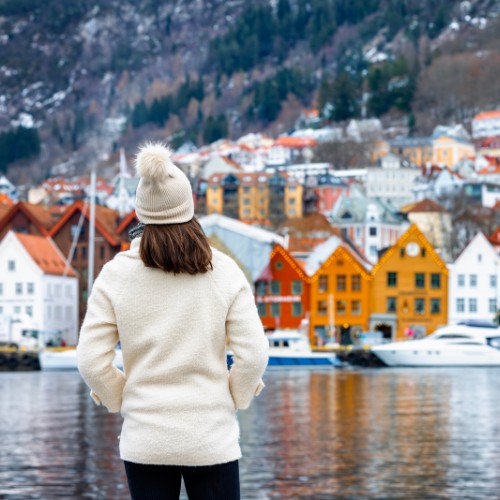 A person in a white winter outfit and beanie stands by a calm body of water, facing colorful houses and boats, with a wooded hillside in the background.