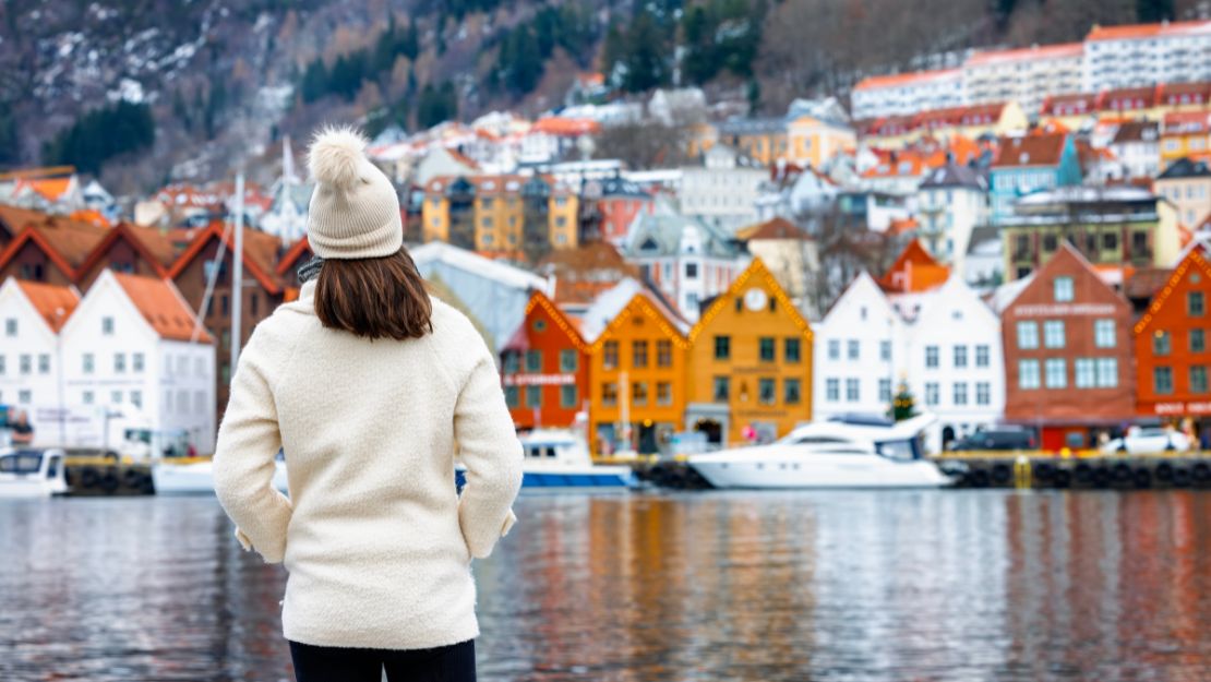 A person in a white winter outfit and beanie stands by a calm body of water, facing colorful houses and boats, with a wooded hillside in the background.