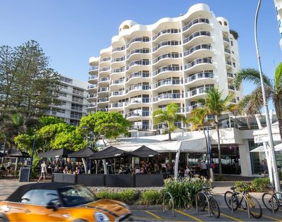 Modern multi-story resort apartment building with curved balconies, palm trees, and street-level shops in Queensland