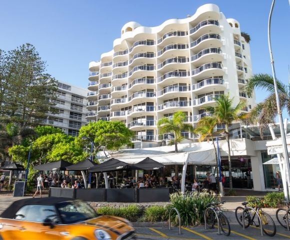 Modern multi-story resort apartment building with curved balconies, palm trees, and street-level shops in Queensland