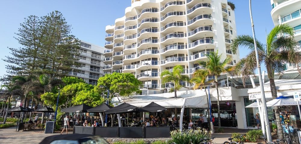 Modern multi-story resort apartment building with curved balconies, palm trees, and street-level shops in Queensland