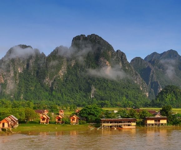 Wooden bungalows and stilted buildings line a muddy river in front of lush green mountains shrouded in clouds.