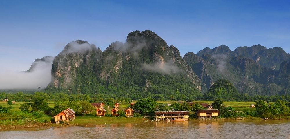 Wooden bungalows and stilted buildings line a muddy river in front of lush green mountains shrouded in clouds.