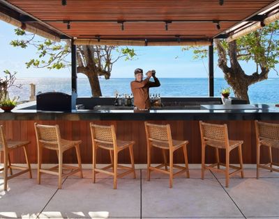 A bartender shakes a cocktail at a modern beachfront bar with a view of the ocean, lined with wooden bar stools.