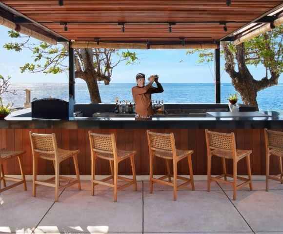 A bartender shakes a cocktail at a modern beachfront bar with a view of the ocean, lined with wooden bar stools.