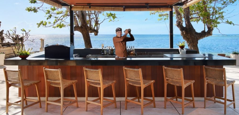 A bartender shakes a cocktail at a modern beachfront bar with a view of the ocean, lined with wooden bar stools.