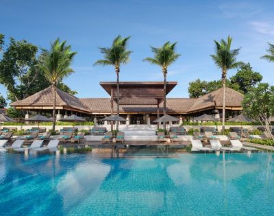 A resort swimming pool with lounge chairs and umbrellas in front of a tropical building with thatched roofs and palm trees.