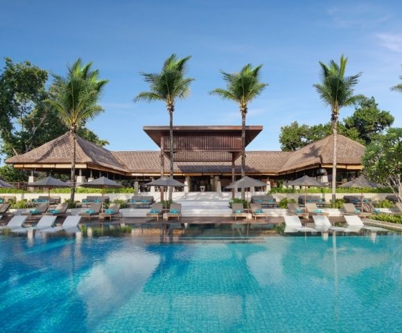 A resort swimming pool with lounge chairs and umbrellas in front of a tropical building with thatched roofs and palm trees.