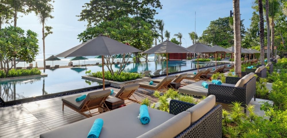 Resort poolside with lounge chairs, umbrellas, and a pool overlooking a beach.