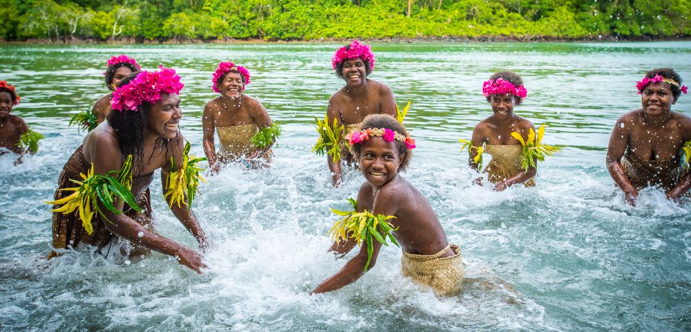 A group of women in traditional attire and pink flower crowns splash in clear water, with lush green foliage in the background.