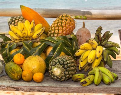 A vibrant, overflowing display of tropical fruits, including bananas, pineapples, oranges, a papaya, and a melon, arranged on a rustic wooden surface with a blurred ocean and bamboo backdrop.