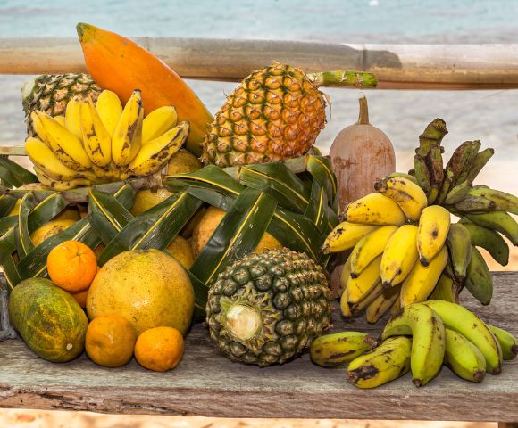 A vibrant, overflowing display of tropical fruits, including bananas, pineapples, oranges, a papaya, and a melon, arranged on a rustic wooden surface with a blurred ocean and bamboo backdrop.
