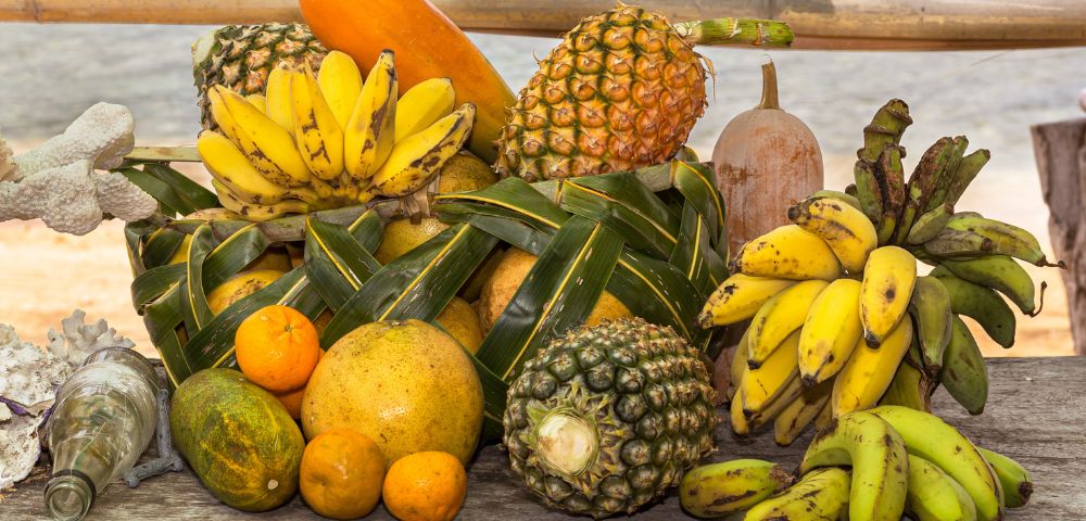 A vibrant, overflowing display of tropical fruits, including bananas, pineapples, oranges, a papaya, and a melon, arranged on a rustic wooden surface with a blurred ocean and bamboo backdrop.