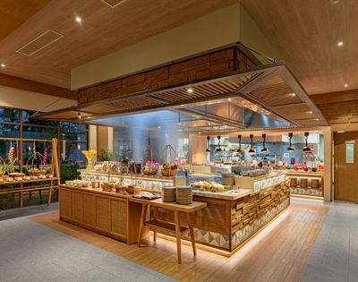 Buffet area in a modern restaurant with wooden decor, food displays, plates, and a visible kitchen in the background.