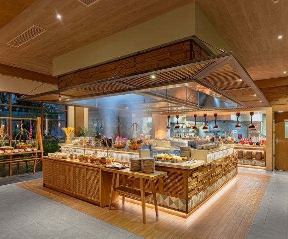 Buffet area in a modern restaurant with wooden decor, food displays, plates, and a visible kitchen in the background.