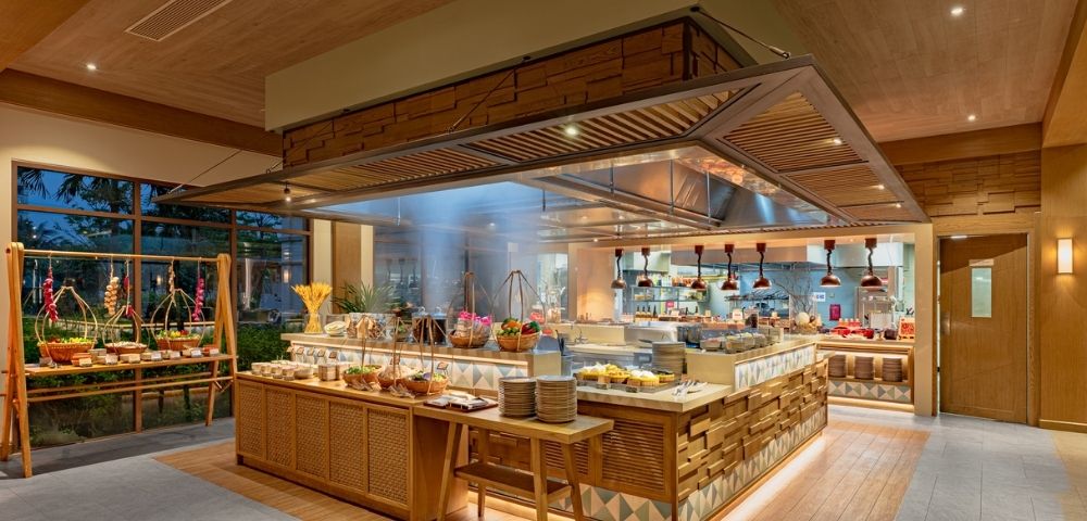 Buffet area in a modern restaurant with wooden decor, food displays, plates, and a visible kitchen in the background.