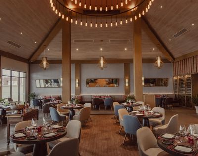 Spacious, modern restaurant interior with wood accents, round tables set for dining, mixed seating, and elegant hanging light fixtures.