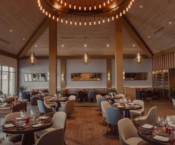 Spacious, modern restaurant interior with wood accents, round tables set for dining, mixed seating, and elegant hanging light fixtures.
