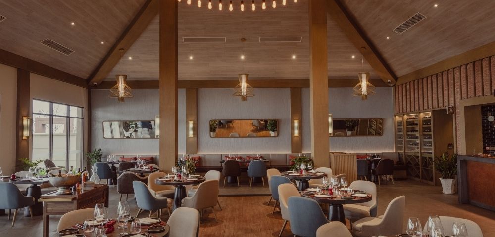 Spacious, modern restaurant interior with wood accents, round tables set for dining, mixed seating, and elegant hanging light fixtures.