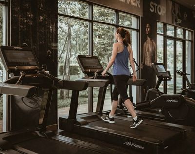 A woman in workout clothes walking on a treadmill in a gym with large windows showing trees outside.