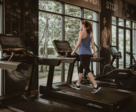 A woman in workout clothes walking on a treadmill in a gym with large windows showing trees outside.