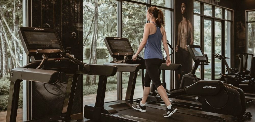 A woman in workout clothes walking on a treadmill in a gym with large windows showing trees outside.