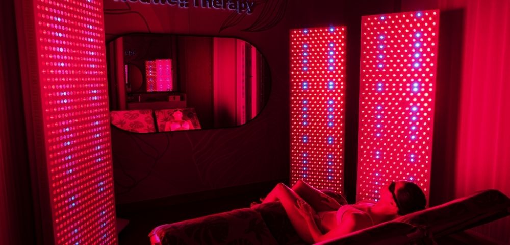 Person relaxing on a lounge chair surrounded by red LED light panels in a dimly lit red therapy room.