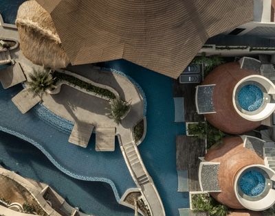 Top-down aerial view of a resort swimming pool with curved walkways, lounge areas, and modern buildings