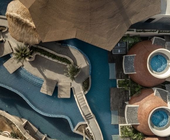 Top-down aerial view of a resort swimming pool with curved walkways, lounge areas, and modern buildings