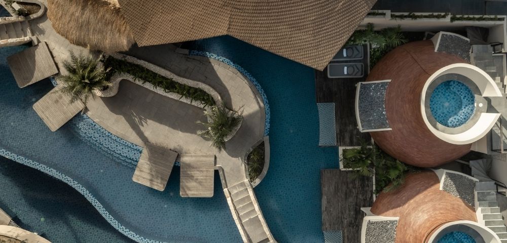 Top-down aerial view of a resort swimming pool with curved walkways, lounge areas, and modern buildings