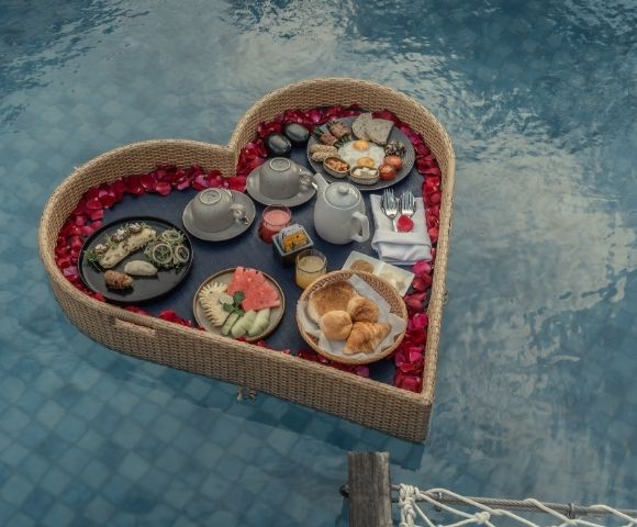 Heart-shaped floating breakfast tray with pastries, fruit, and beverages in a swimming pool